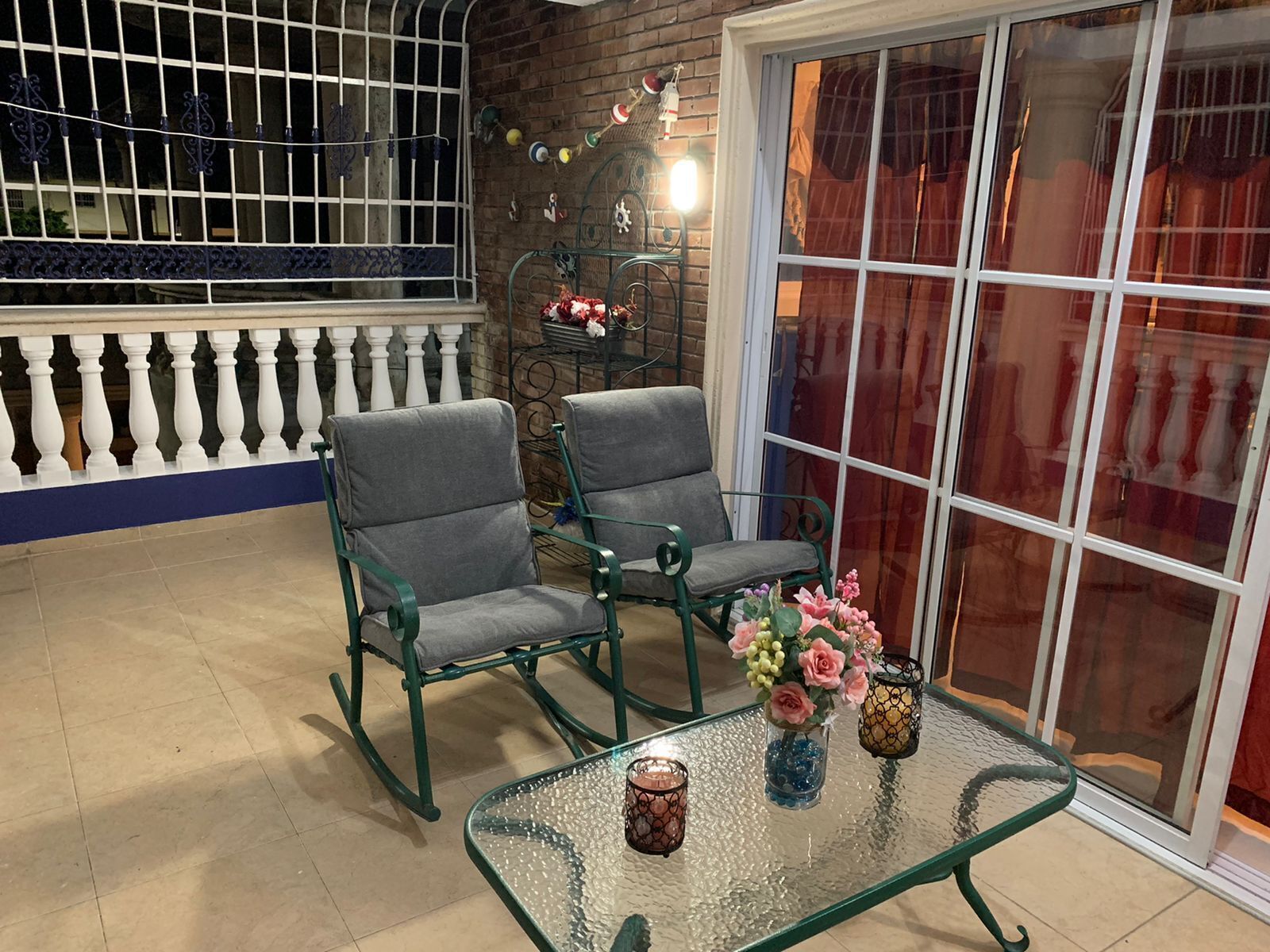 A balcony with two green chairs, a glass table with candles, and a view of a building through a large window.