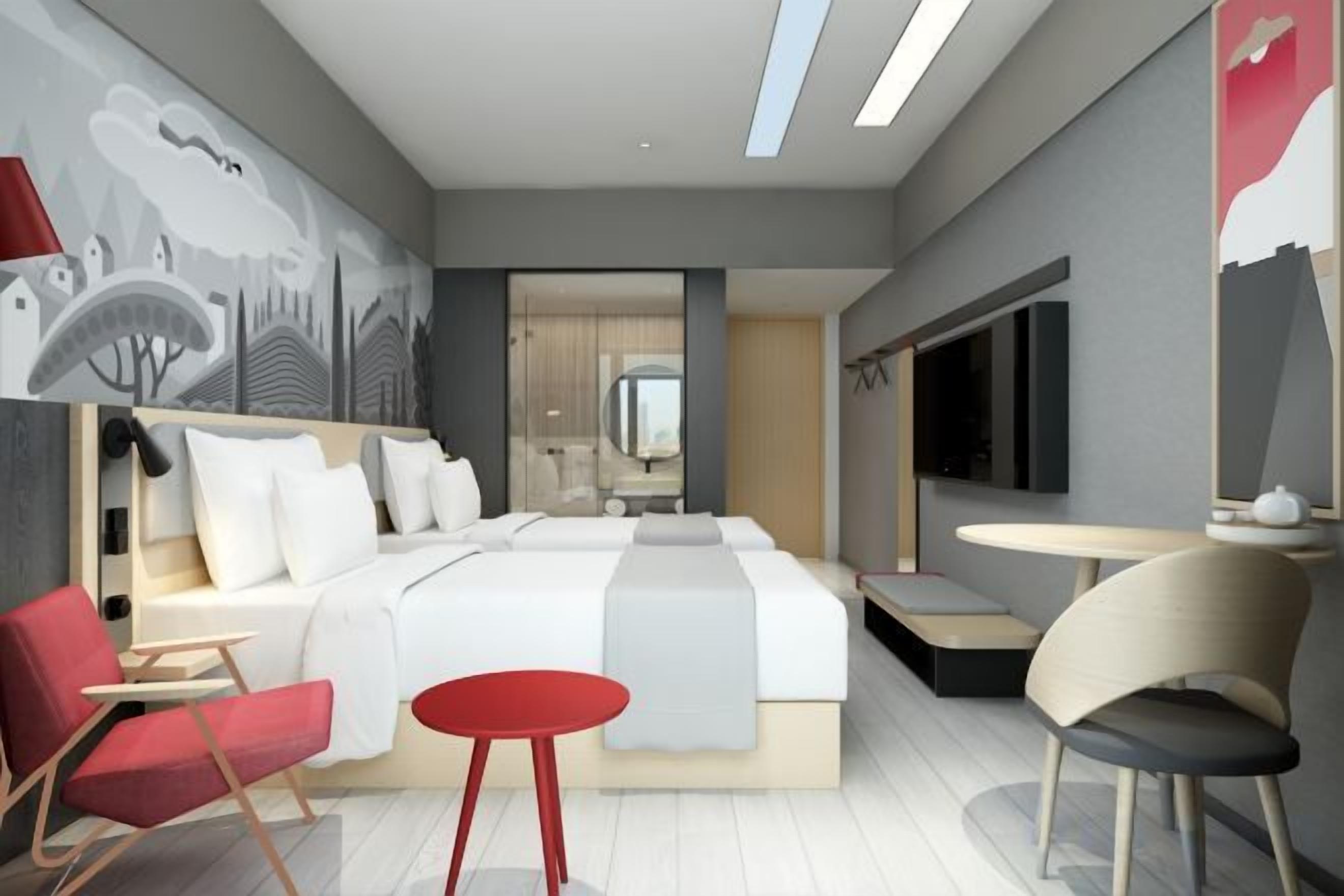 Una habitación de hotel moderna con una cama grande, dos sillas rojas, una mesita redonda y un televisor.