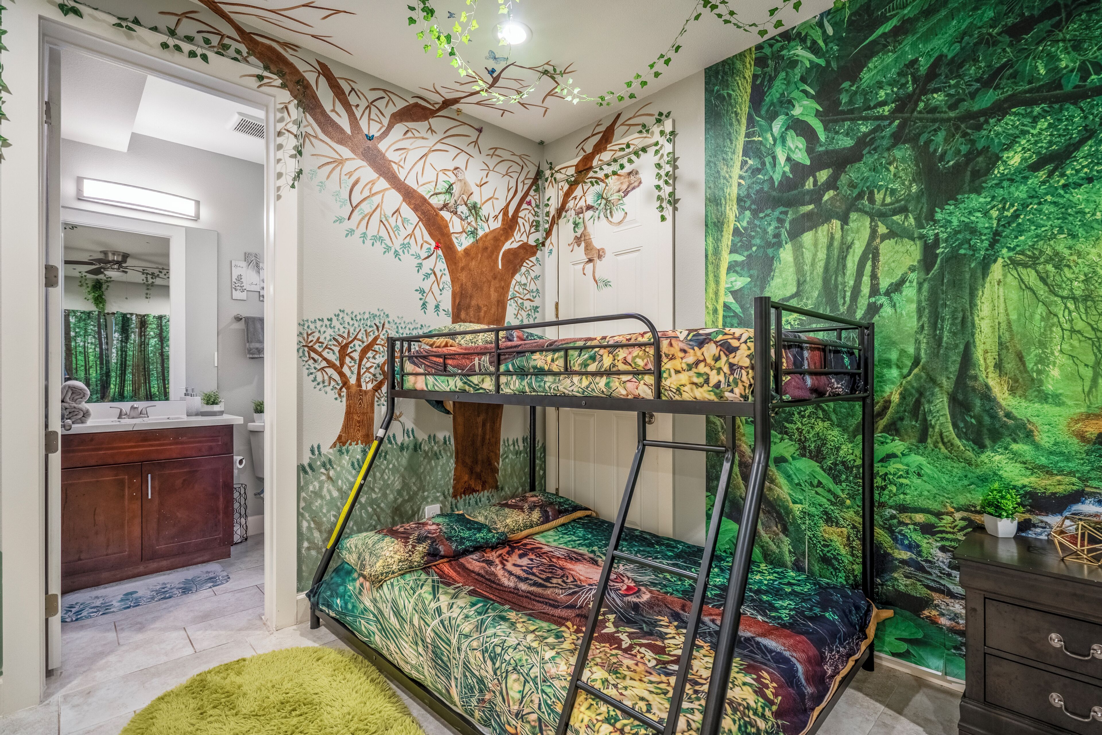 A bedroom with bunk beds, a large forest mural, a vanity with a mirror, and a green rug.