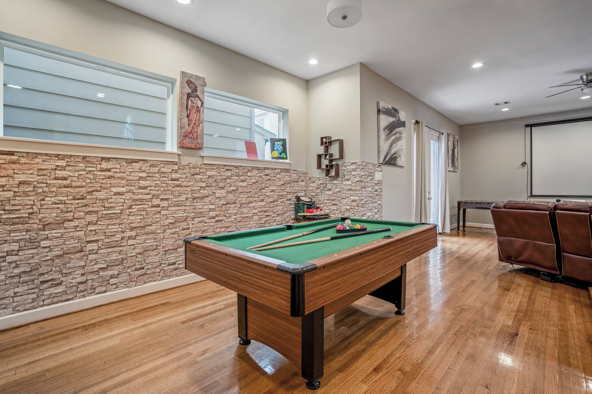 A pool table in a room with wooden flooring, stone wall, and a ceiling fan.