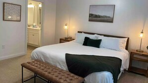 A bedroom with a large bed, a bench, a bedside table, a nightstand, a mirror, and a picture on the wall. - Cheerful Three-Bedroom Residential Home (Richmond)