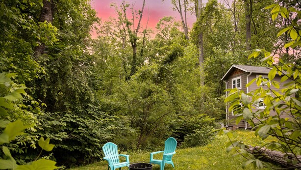 A rustic outdoor setting with two turquoise Adirondack chairs around a campfire, surrounded by lush greenery and a small cabin in the background.