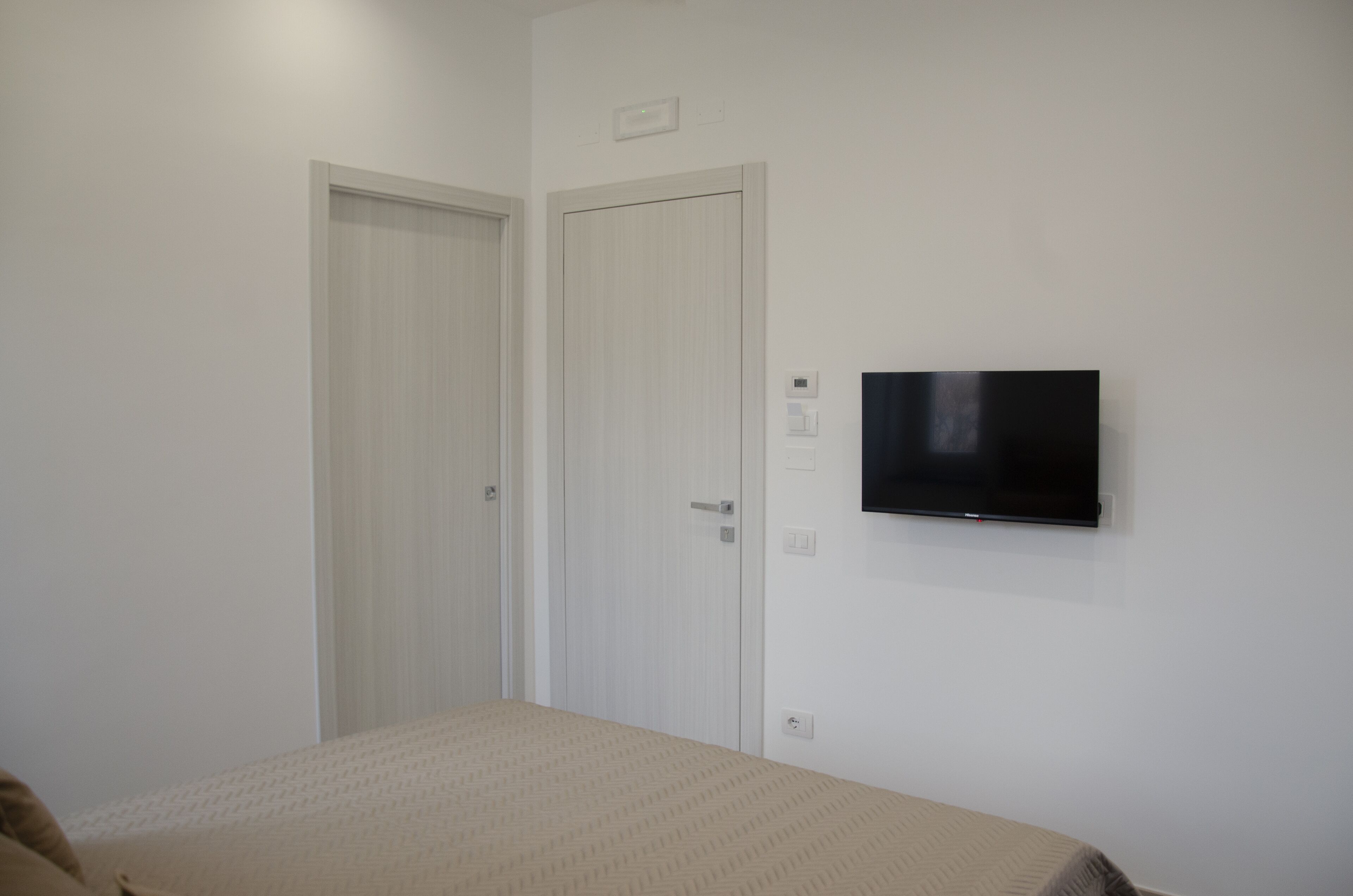 A bedroom with a bed, two white doors, a mounted TV, and a light switch.