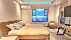 Quarto com uma cama grande, janela com vista para o mar, uma cadeira e escrivaninha com televisão.