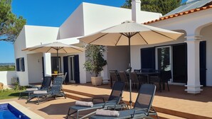 A modern outdoor pool area with lounge chairs and umbrellas, a dining area with a table and chairs, and a view of the surrounding landscape.
