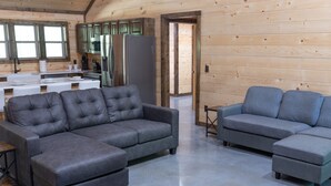 Una sala moderna con dos sofás grises y una mesa de centro, paredes de madera y una zona de cocina con refrigerador y armarios.