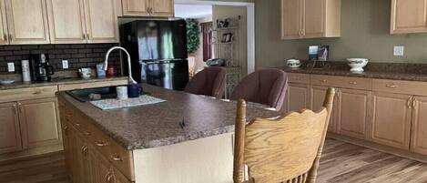 A kitchen with wooden cabinets, a stainless steel refrigerator, a granite countertop, and a wooden chair.
