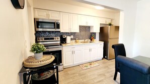 Cocina compacta con armarios blancos, refrigerador de acero inoxidable, microondas y cafetera. Hay un pequeño comedor con una mesa y sillas.
