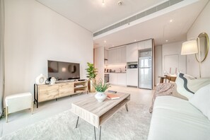 Una sala de estar moderna con una mesa de centro de madera, un sofá y un televisor. Se observa una cocina al fondo con armarios blancos y un refrigerador.