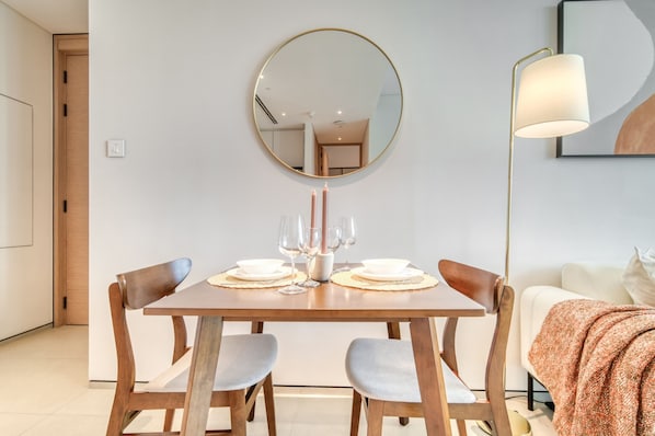 A dining area with a wooden table set for two, a round mirror above, and a modern floor lamp to the right.