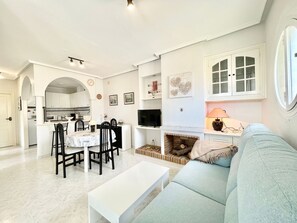 A modern living room with a sofa, a dining table with chairs, a fireplace, and a kitchen in the background.