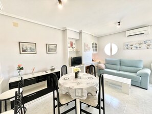 A modern living room with a light blue sofa, a dining area with a table and chairs, and a TV on a stand.
