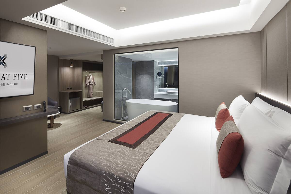 A modern hotel room with a large bed, a bathroom with a bathtub, a shower, and a vanity. There is a desk with a chair and a large window.