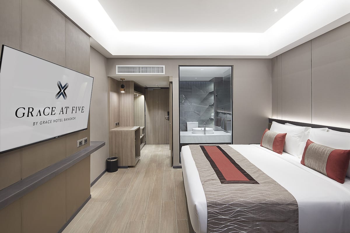 A modern hotel room with a large bed, a TV displaying the hotel's name, a bathroom with a glass shower, and a small seating area.