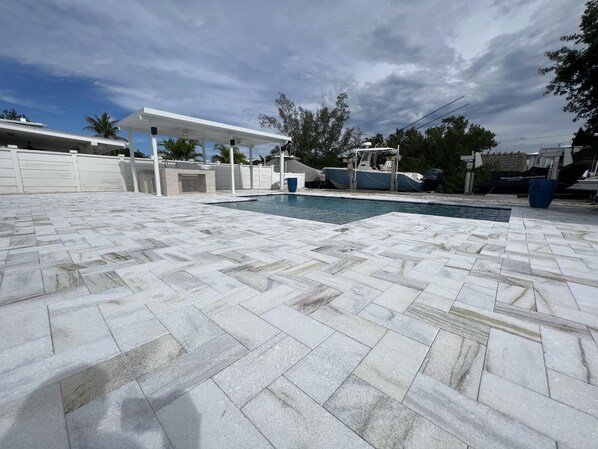A swimming pool with a tiled deck, a covered seating area, and a boat docked nearby.