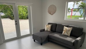A modern living room with a grey sectional sofa, two decorative pillows, a round wall hanging, and a large sliding glass door leading to a patio area. - Great House in Key Largo (Key Largo)