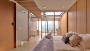 A modern bedroom with a large bed, a glass partition, and a view of a balcony with outdoor furniture.
