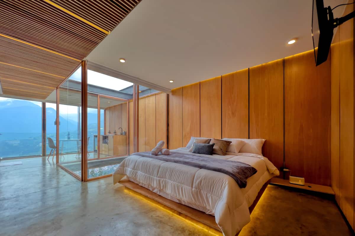 Une chambre moderne avec un grand lit, des murs recouverts de panneaux en bois et une porte en verre donnant sur un balcon avec vue sur la ville.