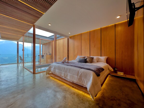 A modern bedroom with a large bed, wooden panel walls, and a glass door leading to a balcony with a city view. - Deluxe Volcán (Baños de Agua Santa)