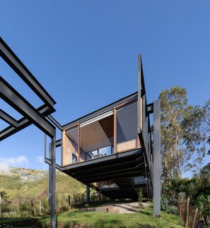 A modern house with a large glass balcony, surrounded by greenery and hills. - Deluxe Volcán (Baños de Agua Santa)