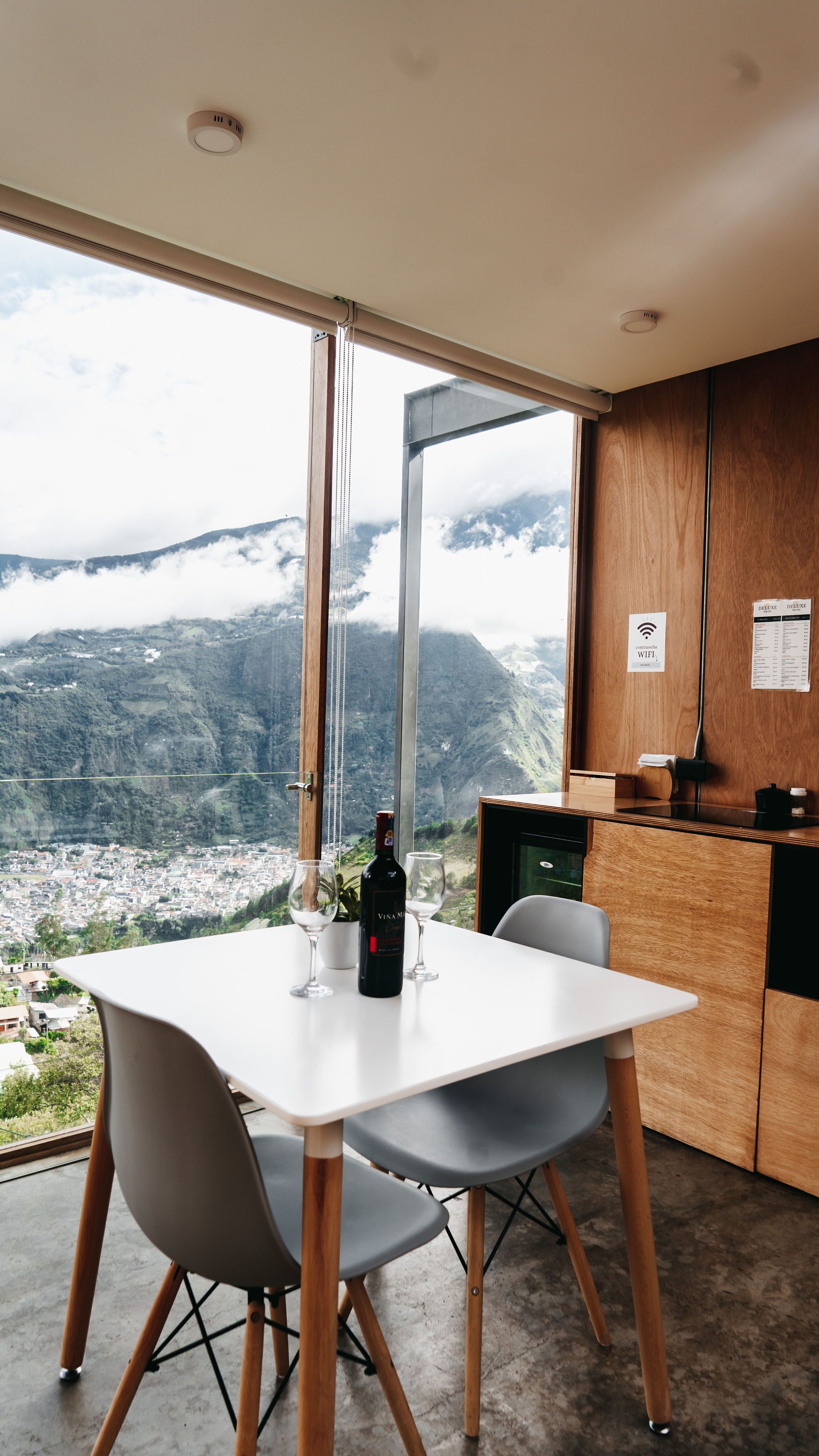 Uma área de refeições com mesa e cadeiras, uma garrafa de vinho e vista para uma paisagem montanhosa através de grandes janelas.