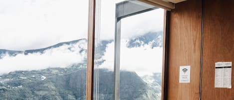 Uma área de refeições com mesa e cadeiras, uma garrafa de vinho e vista para uma paisagem montanhosa através de grandes janelas.