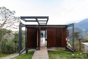 Una estructura moderna de madera con puerta de cristal, un pasillo de madera que la conduce y una vista de montañas al fondo.