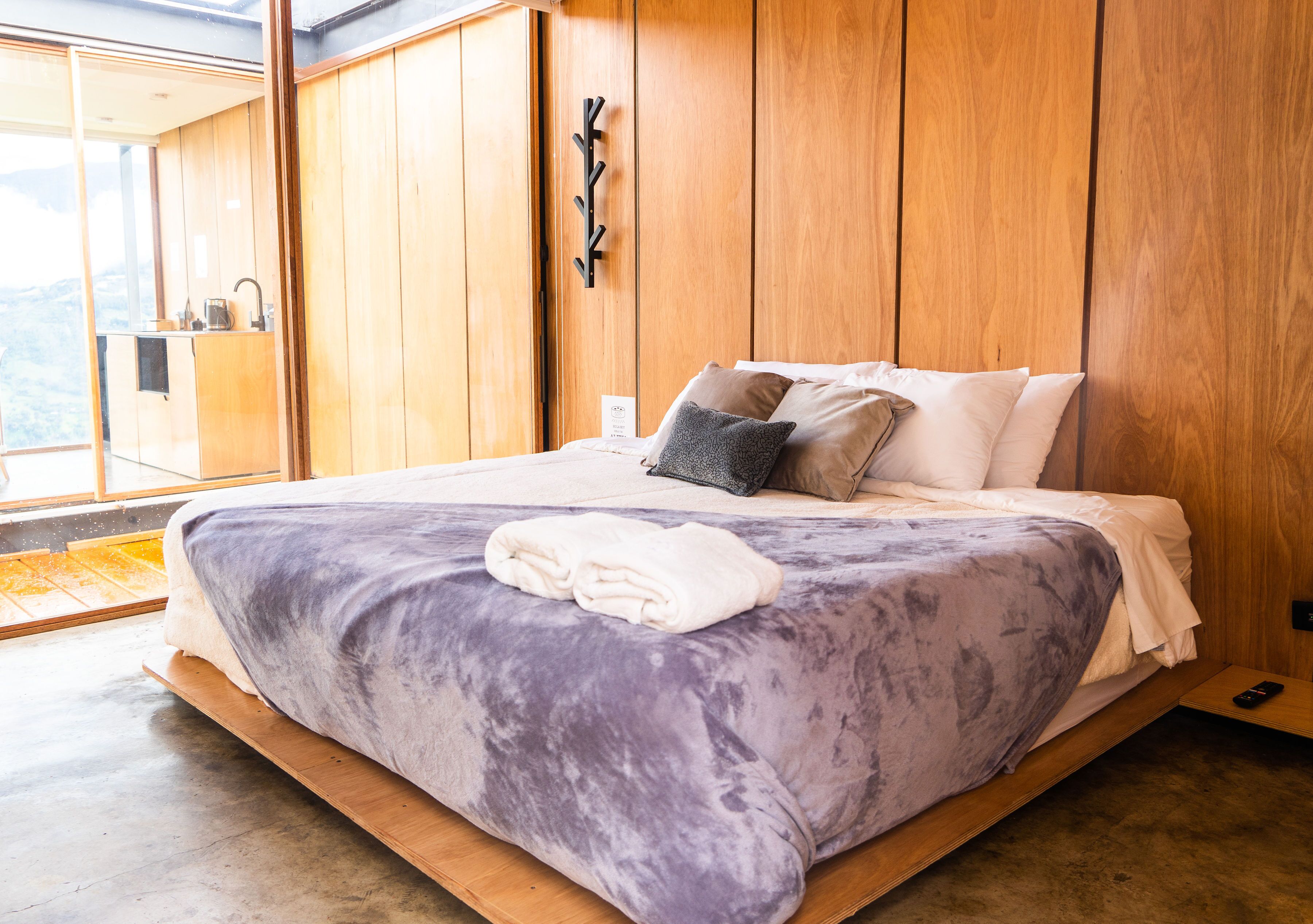 A bedroom with a large bed, wooden walls, a small bedside table, and a view of the outdoors.