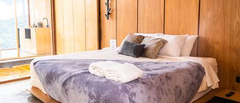 A bedroom with a large bed, wooden walls, a small bedside table, and a view of the outdoors.