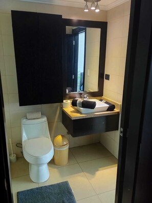 A modern bathroom with a white toilet, a sink with a towel, and a mirror above it. - Departamento Emporium (Guayaquil)