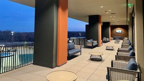 An outdoor seating area with high chairs and tables, a pool in the background, and a TV screen mounted on the wall.