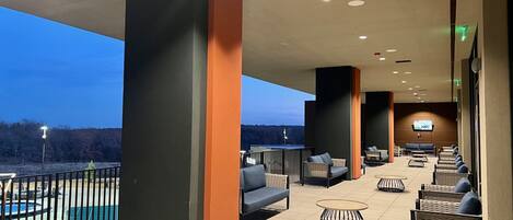 An outdoor seating area with high chairs and tables, a pool in the background, and a TV screen mounted on the wall.