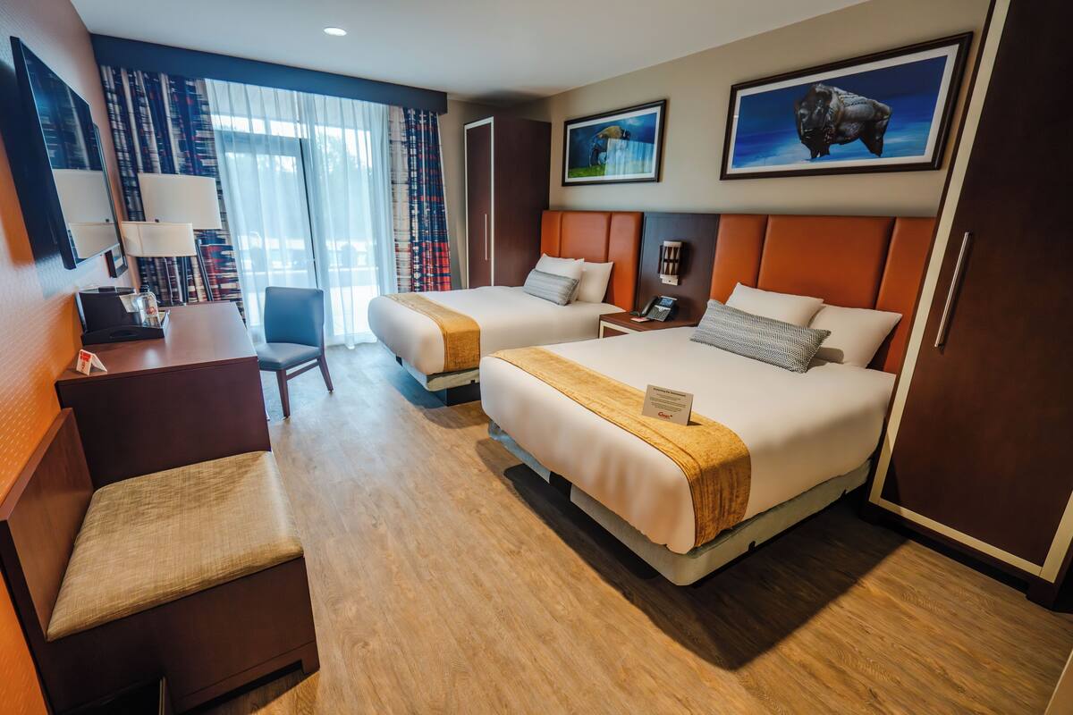 A hotel room with two beds, a desk, a chair, a TV, and a large window with curtains.