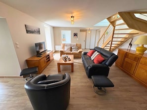 A living room with a sofa, coffee table, and a black leather chair. There is a wooden cabinet, a television, and a staircase with a wooden railing.