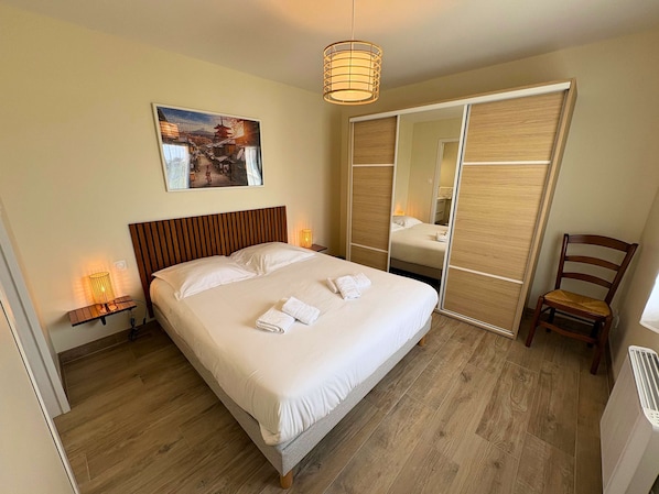 A neatly made bed with white linens, a wooden headboard, a bedside lamp, a wooden floor, a chair, and a large mirror.