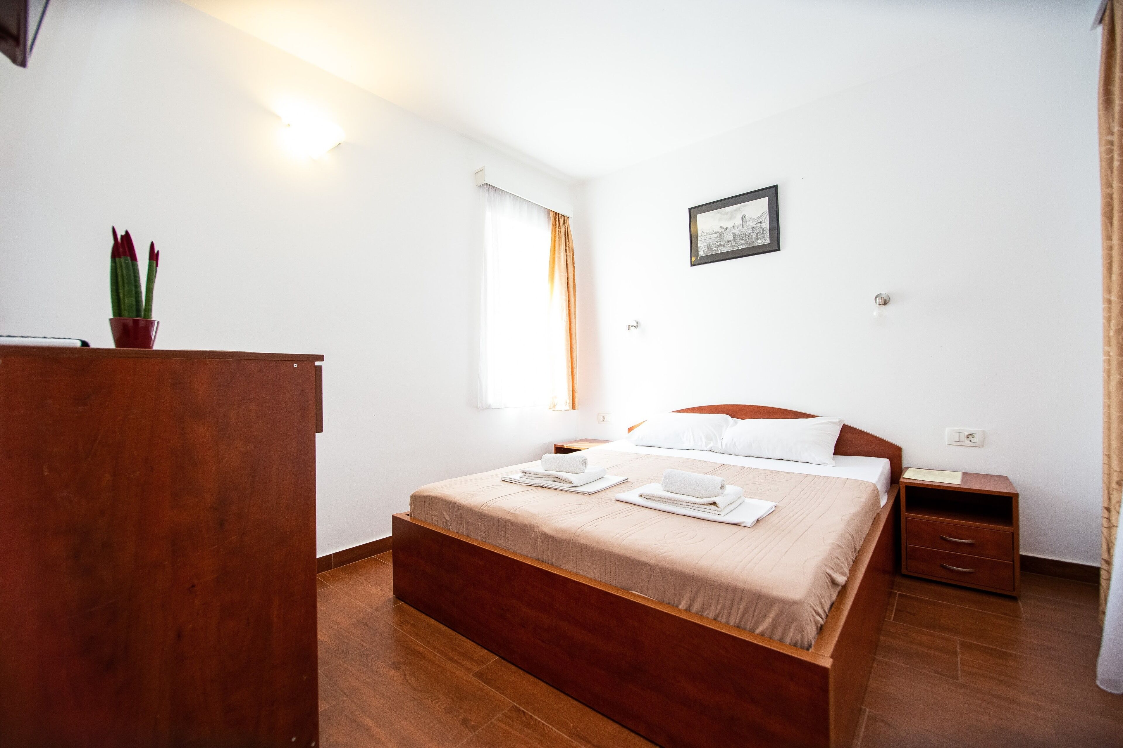 Quarto arrumado com cama de madeira, mesa de cabeceira, criadeira e uma pequena planta em vaso.