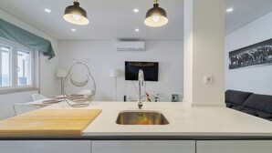A modern kitchen with a central island, a built-in sink, overhead lighting, a flat-screen TV, and a large window. - Fonte Nova Nest (Sesimbra)