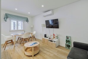 Uma sala de estar moderna com televisão de tela plana, um espaço de refeições com cadeiras, uma mesa de centro redonda, um sofá e um pequeno armário com objetos decorativos.