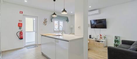 Una cocina moderna con una isla central, iluminación colgante, un televisor de pantalla plana y un sofá gris en una sala de estar.