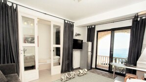 Een moderne hotelkamer met een bed, een sofa, een televisie, een koelkast en een balkon met uitzicht op zee.