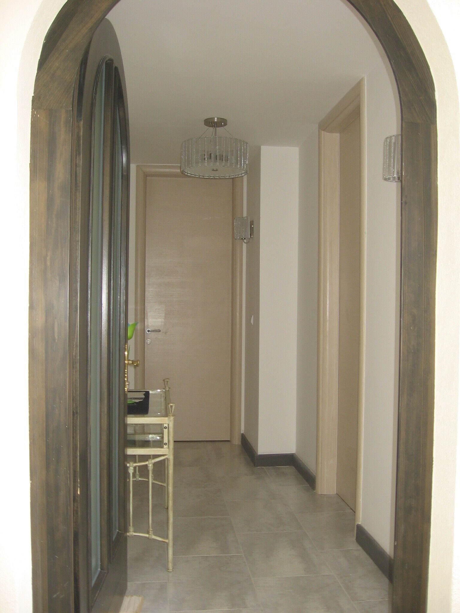 Un couloir avec une porte en bois, une petite table sur laquelle est posé un vase, et un lustre.