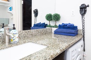 Baño con encimera de granito, lavamanos, secador de pelo, lavadora y dos pequeñas macetas.
