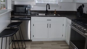 A kitchen with white cabinets, a black microwave, a sink, and a stove. There are three stools and a window with blinds. - home sweet home (Rex)