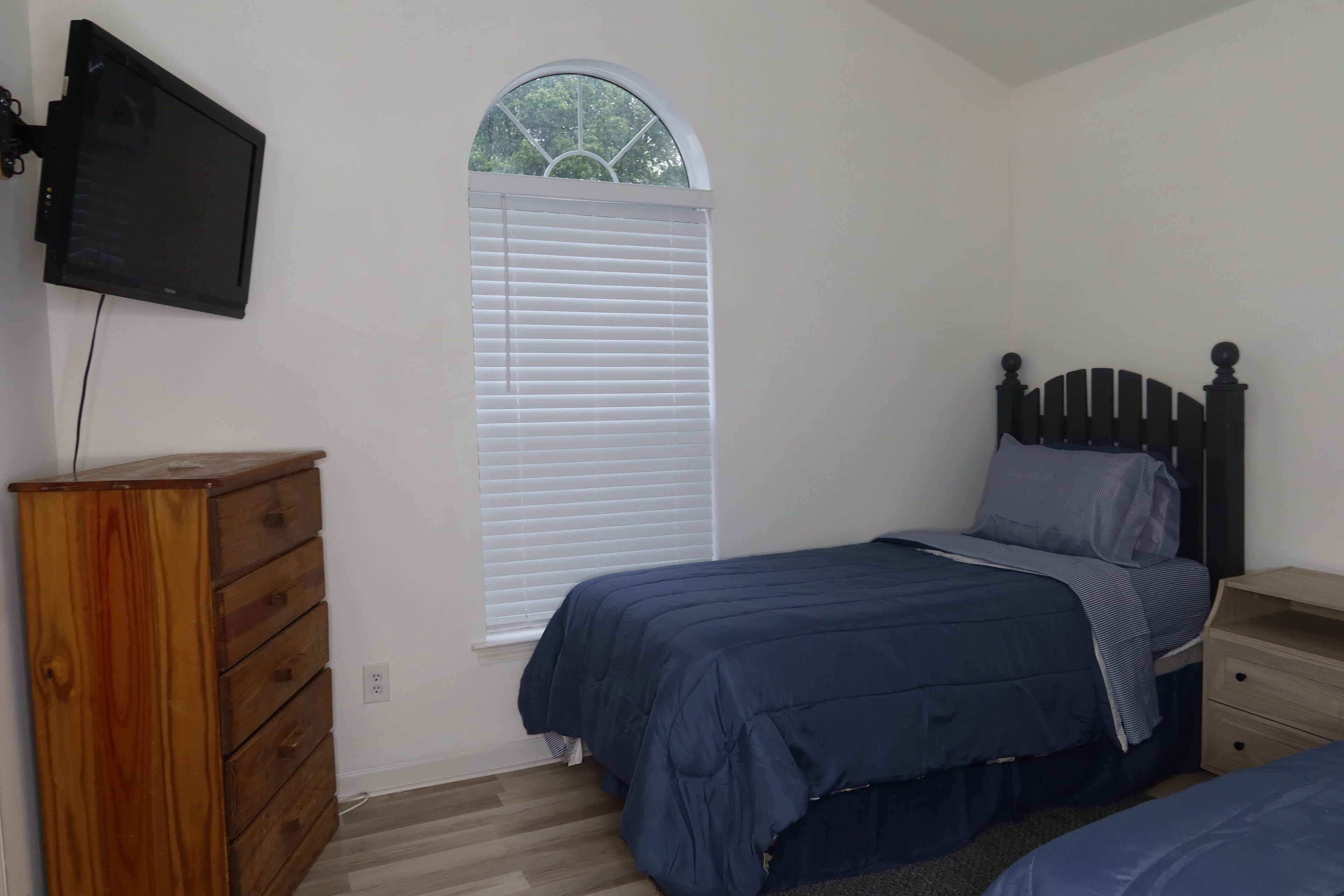 A bedroom with a bed, a wooden dresser, a nightstand, and a TV mounted on the wall.