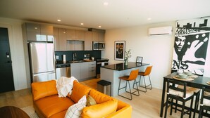 Una cocina moderna con electrodomésticos de acero inoxidable, un comedor con una mesa y sillas negras, y una sala de estar con un sofá naranja.