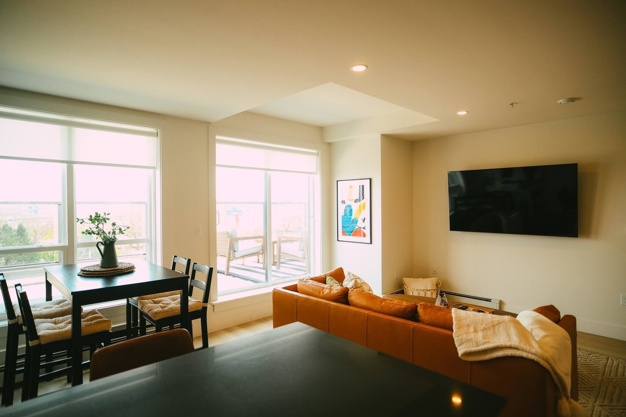 Een moderne woonkamer met een eethoek, grote ramen, een flatscreen televisie en een bank.