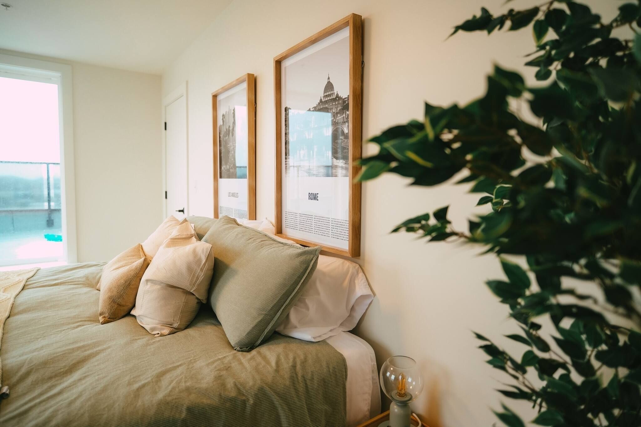 A bedroom with a bed, two framed pictures on the wall, a bedside table with a lamp, and a potted plant.
