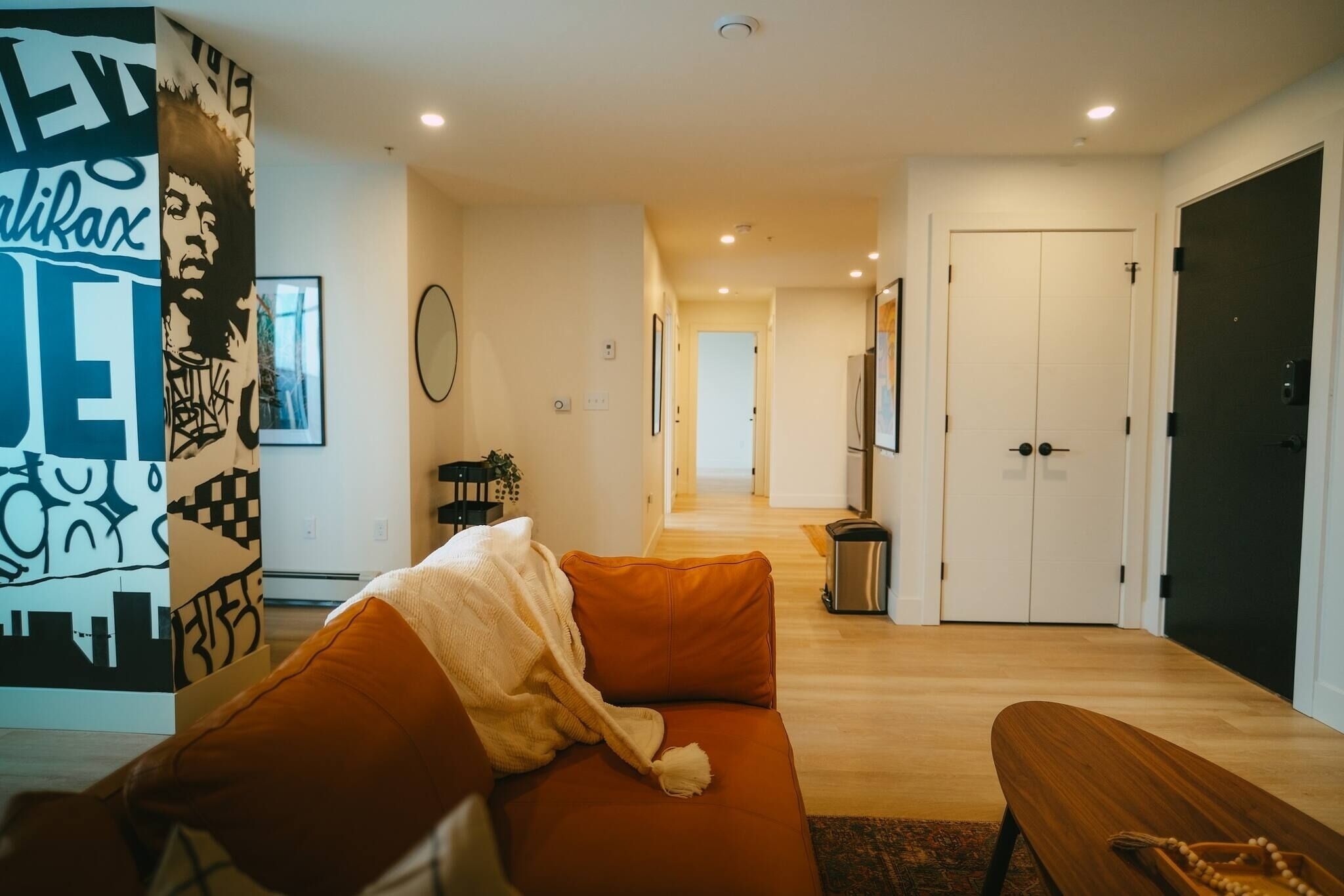 A modern living room with a sofa, a bench, a wall with artwork, a hallway leading to a door, and a trash bin.
