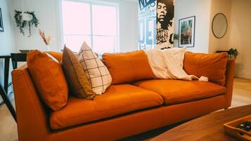 A modern living room with a large orange sofa, a wooden coffee table, and a large window providing natural light.