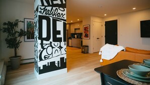 A modern living room with a large black and white artwork, a dining area with a table set for two, and a kitchen in the background. - Chic Cozy Retreat - 2BR - Stunning North End Views (Halifax)